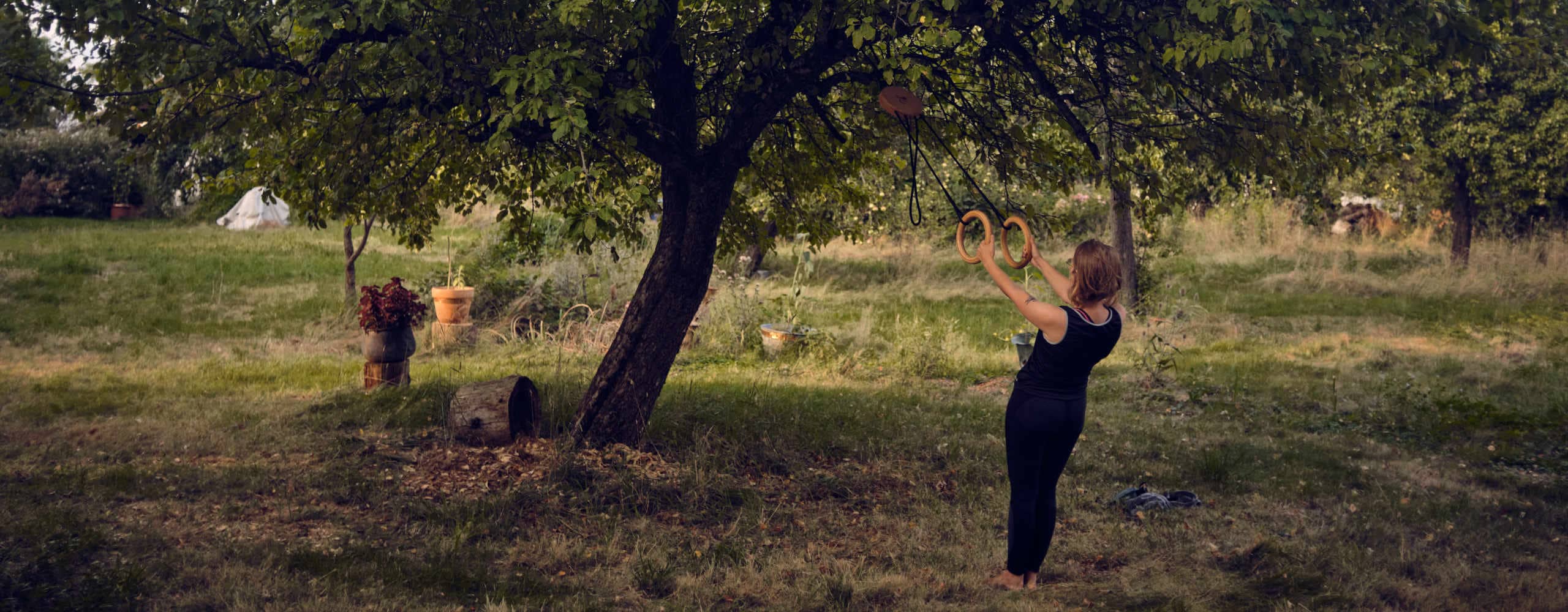 Ein breites Outdoor-Banner zeigt eine Frau, die barfuß im idyllischen Garten an rollholz Turnringen aus Holz trainiert, welche an einem großen Baum befestigt sind.