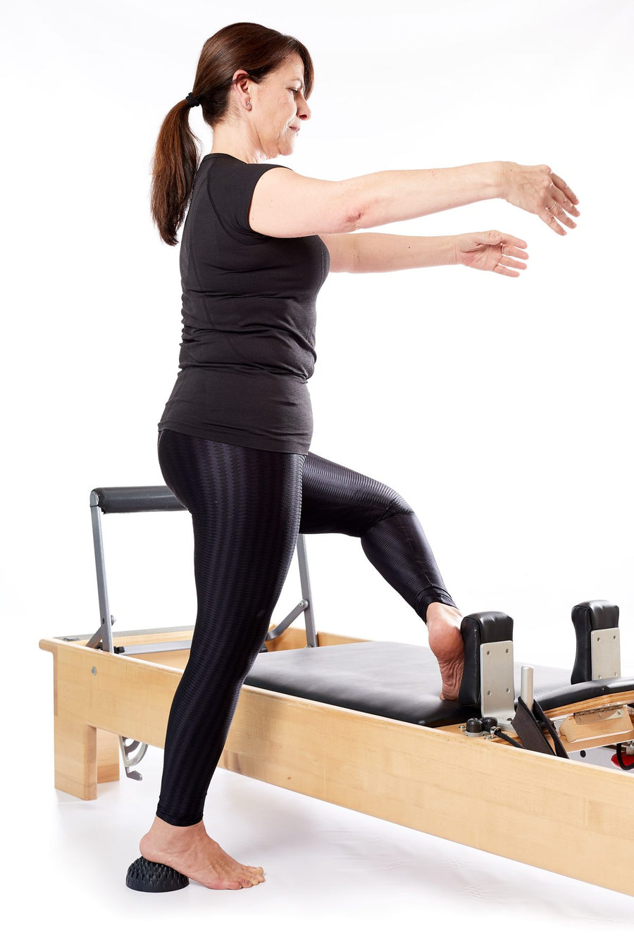 Femme debout à côté d'un reformer de Pilates, un pied sur l'appareil, l'autre sur un grand dôme Lotus