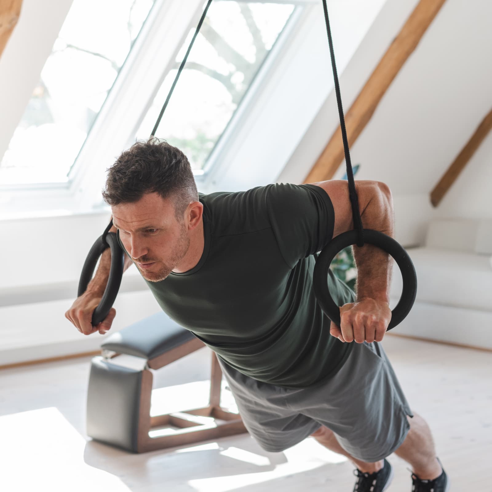 Ein Mann führt Liegestütze (Chest Press) mit dem NOHRD Schlingentrainer Sling an den Holzringen in einem hellen Dachgeschoss-Studio aus.