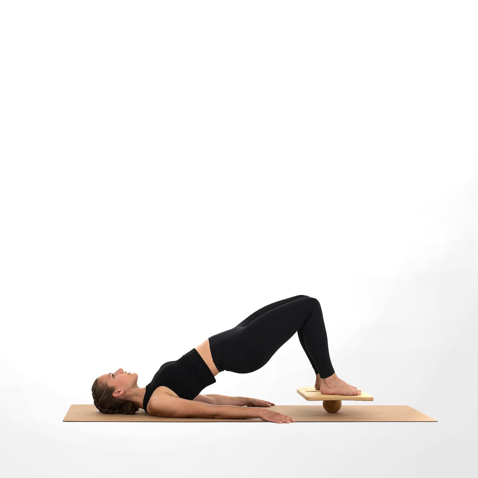 Eine Frau in schwarzer Sportkleidung führt auf einer Korkmatte die Glute-Bridge-Übung aus, indem sie ihre Füße barfuß auf dem rollholz Balanceboard stabilisiert.