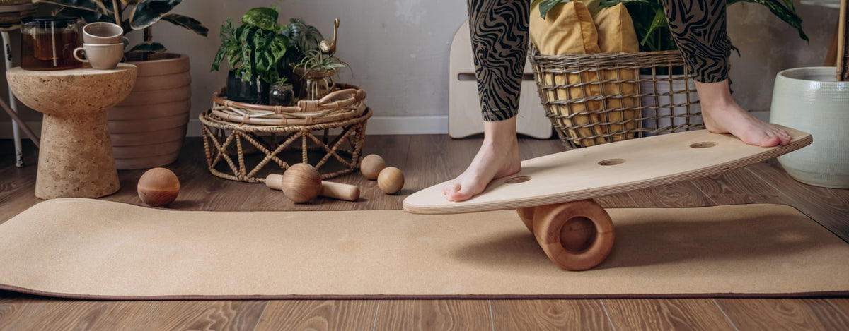 A wide indoor banner shows a wooden rollholz balance board being used barefoot on a cork mat in a cosy living room decorated with plants.