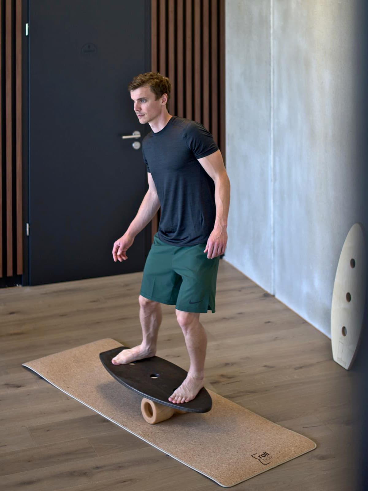 Ein Mann trainiert barfuß im aufrechten Stand auf dem dunklen rollholz Balanceboard aus Holz, das auf einer Korkmatte in einem modern gestalteten Raum liegt.