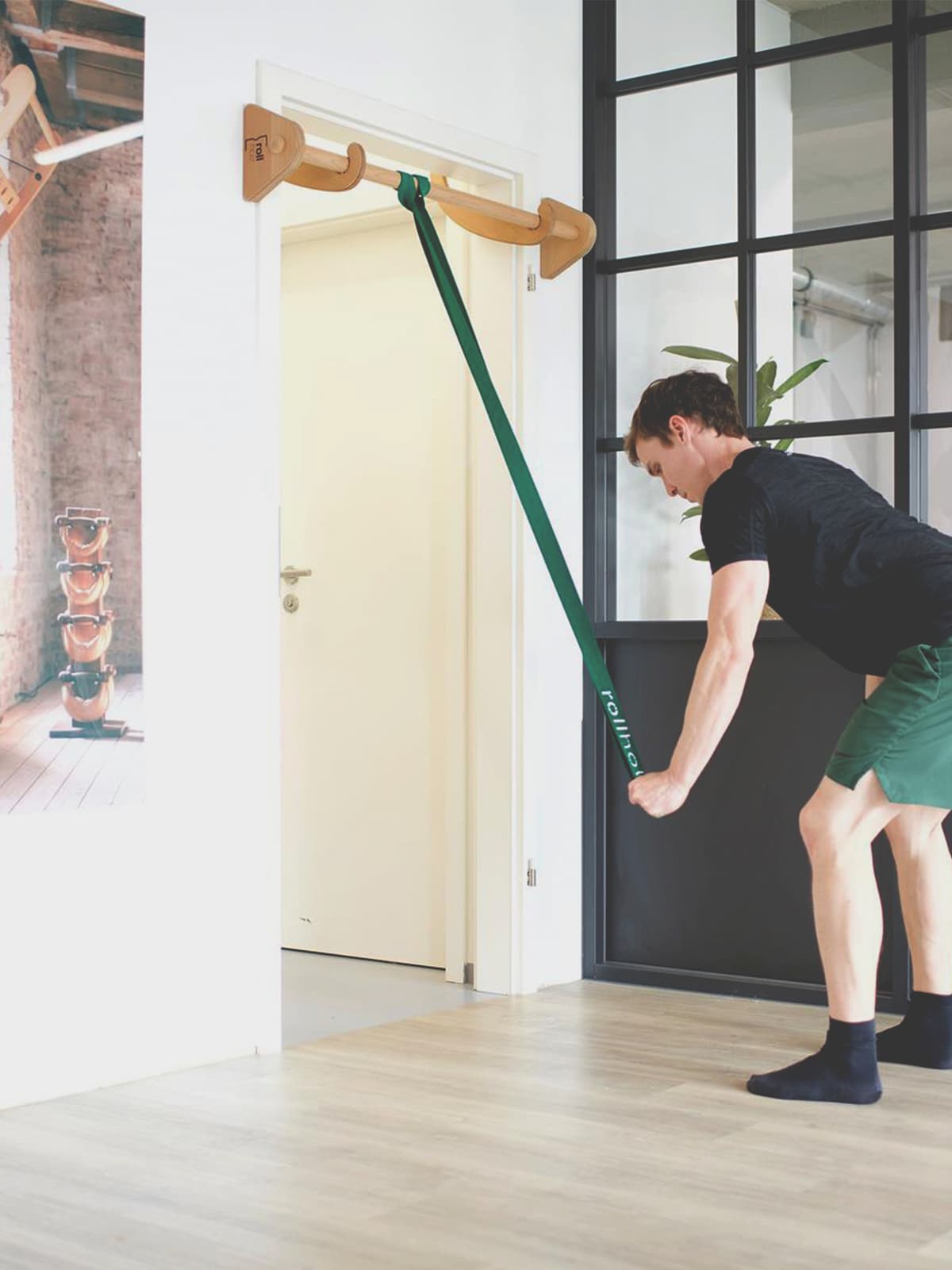 Ein Mann in Sportkleidung nutzt ein grünes rollholz Widerstandsband, das an der Holz-Klimmzugstange im Türrahmen befestigt ist, für eine vorgebeugte Zugübung.