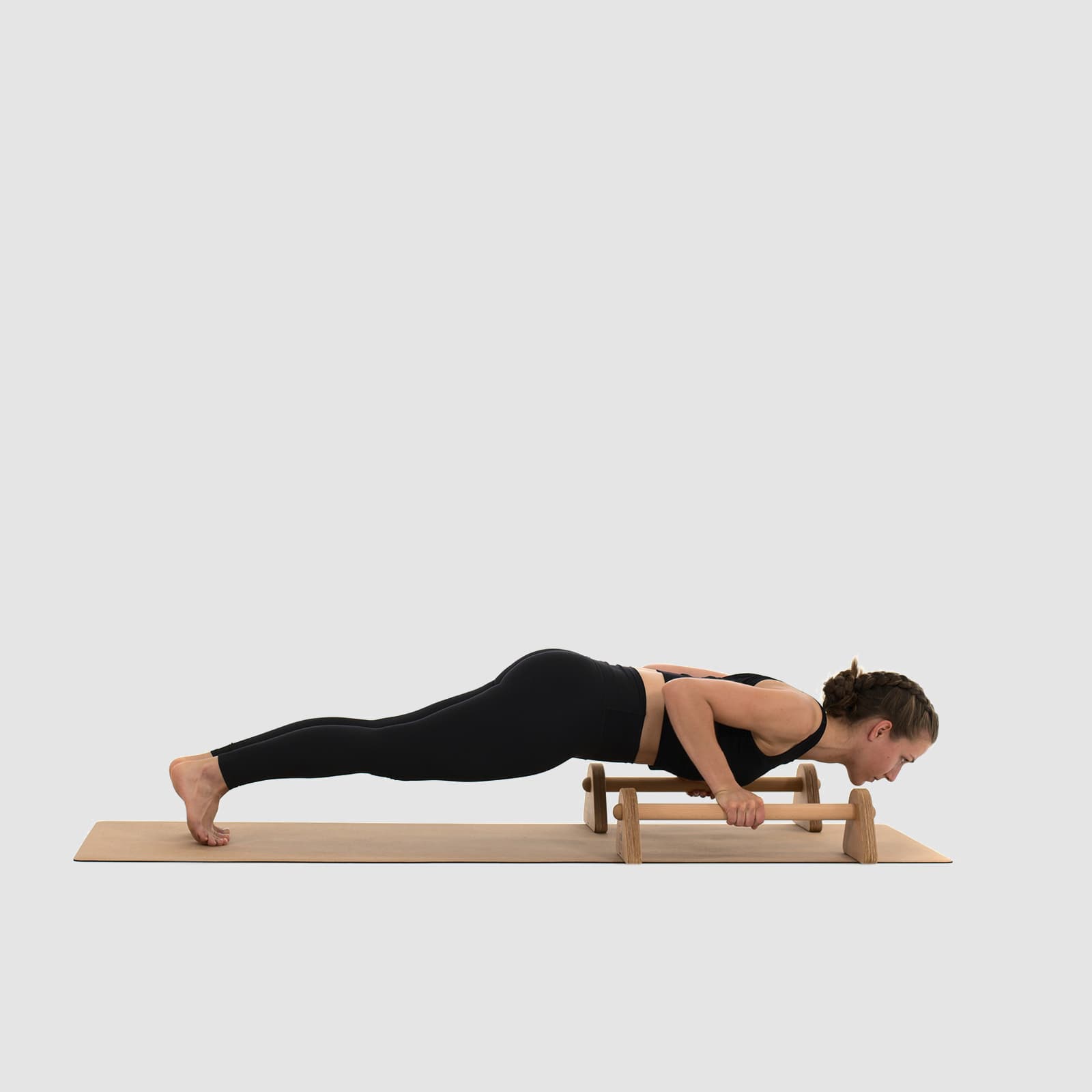 Eine Athletin in schwarzer Sportkleidung führt auf einer Korkmatte tiefe Liegestütze (Push-Ups) an den rollholz Minibarren aus hellem Holz aus.