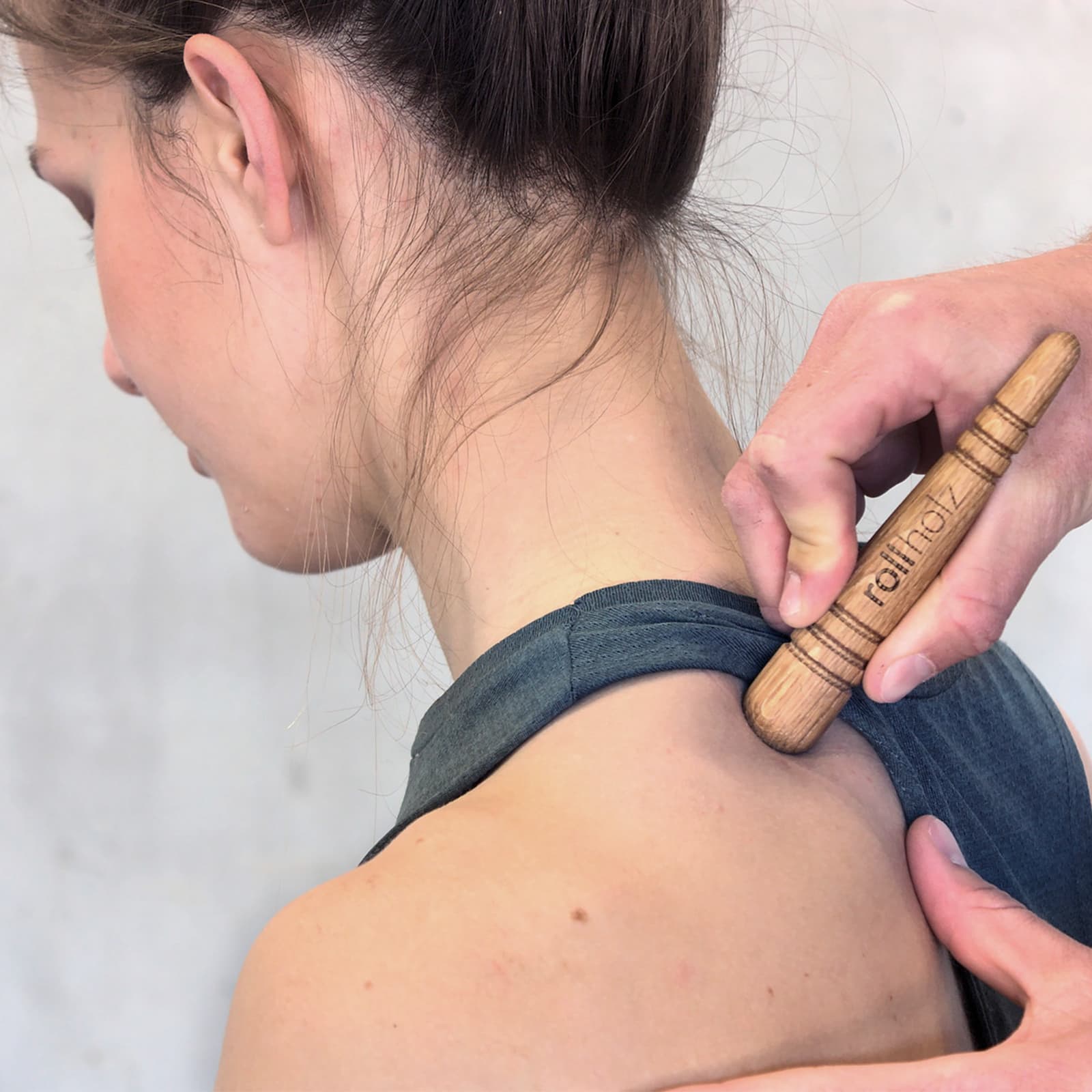 Nahaufnahme einer Anwendung, bei der eine Hand den rollholz Triggerstift aus Holz mit Markenlogo nutzt, um verhärtete Muskeln im Schulter- und Nackenbereich einer Frau zu behandeln.