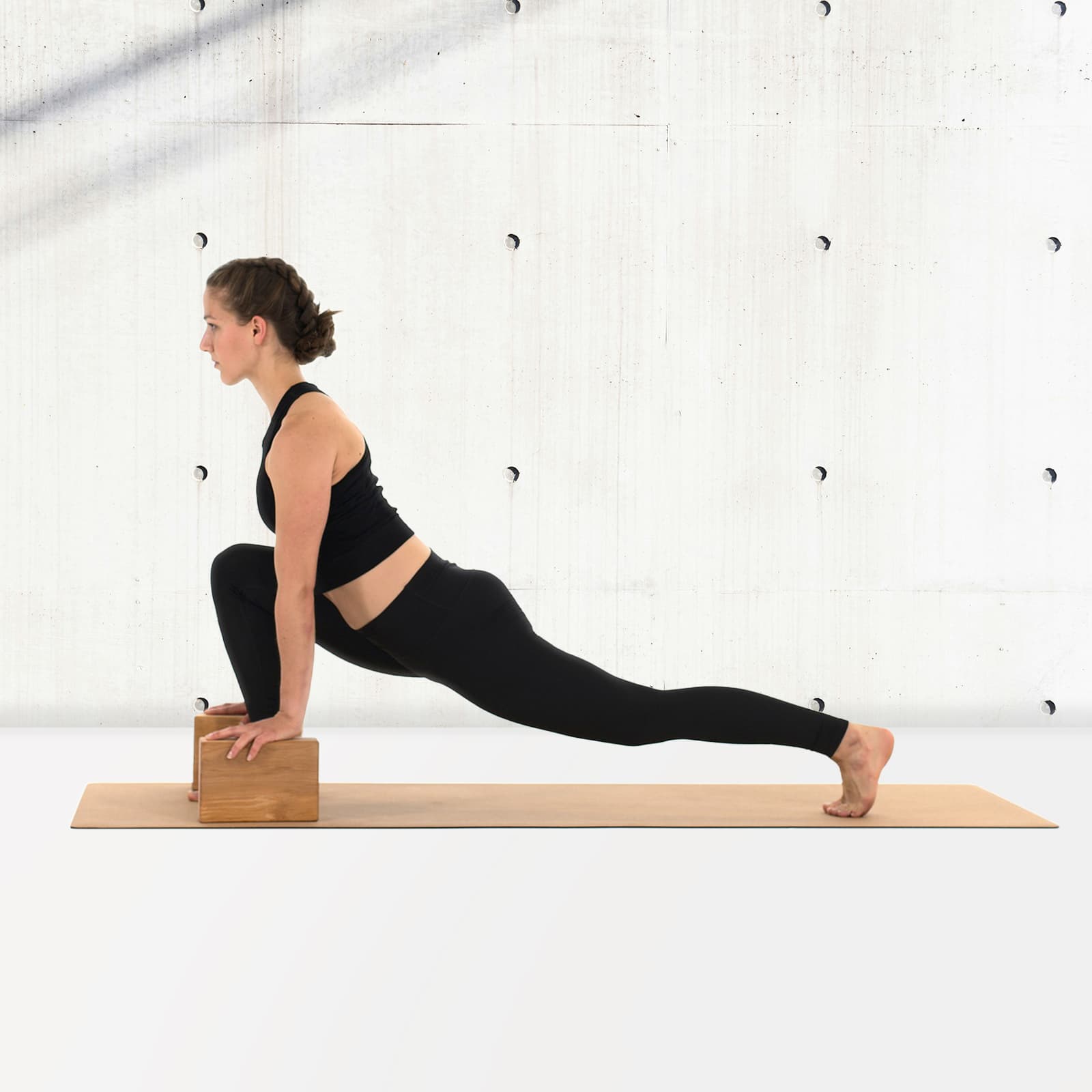 Eine Frau in schwarzer Sportkleidung führt auf einer Korkmatte einen tiefen Ausfallschritt aus und stützt ihre Hände auf zwei rollholz Yogablöcken aus Erlenholz ab, vor einer hellen Sichtbetonwand.