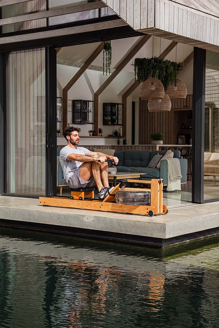 Trainieren mit WaterRower Rudergerät auf Terasse, überdacht, Wasser im Vordergrund.