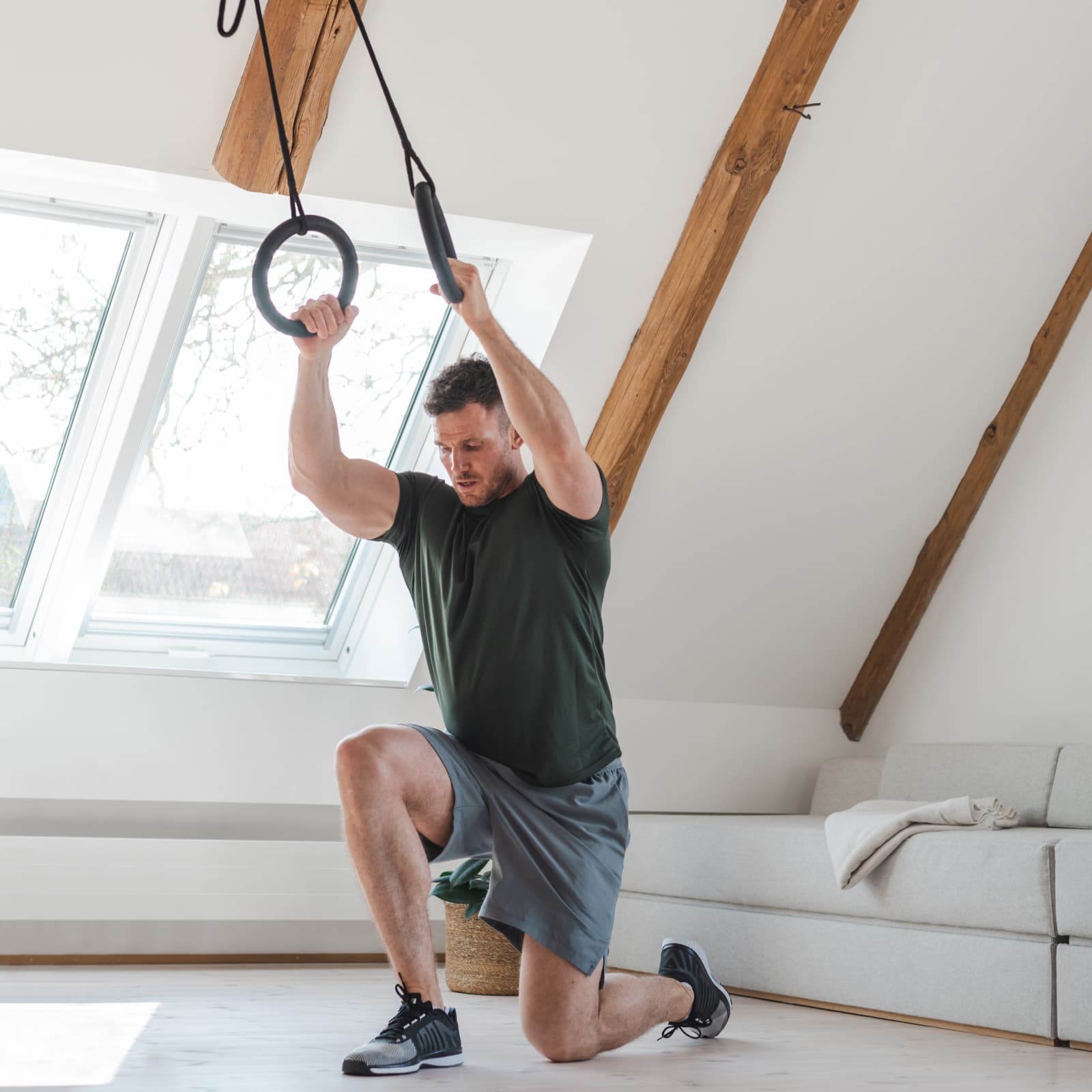 Ein Mann trainiert mit dem NOHRD Schlingentrainer Sling in einem hellen Dachgeschoss-Studio und führt gestützte Ausfallschritte an den Holzringen aus.