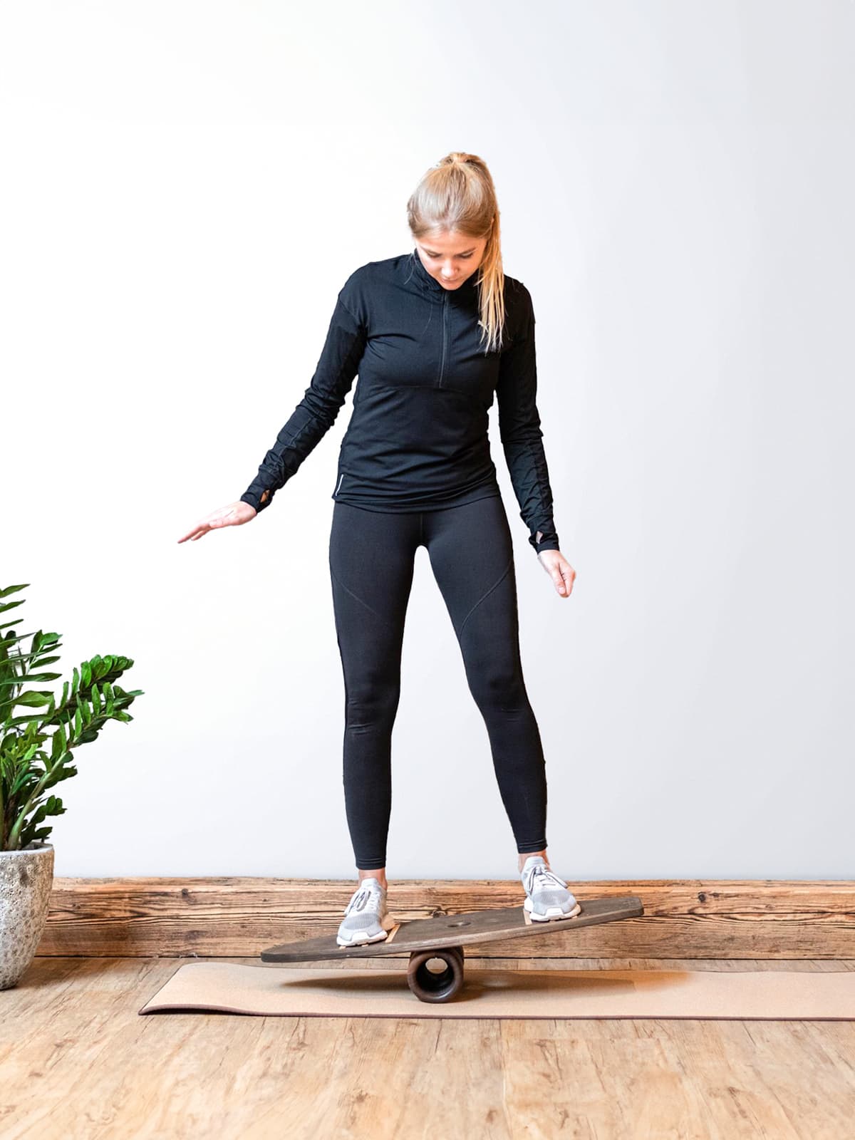 Eine Frau in schwarzer Sportkleidung trainiert fokussiert im aufrechten Stand auf dem dunklen rollholz Balanceboard aus Holz, das auf einer Korkmatte liegt.