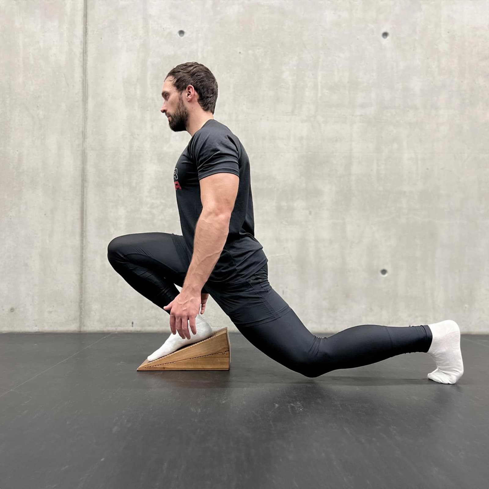 Ein Mann in Sportkleidung führt auf Socken einen tiefen Ausfallschritt aus, wobei der vordere Fuß erhöht auf dem rollholz Balancekeil aus Holz platziert ist.
