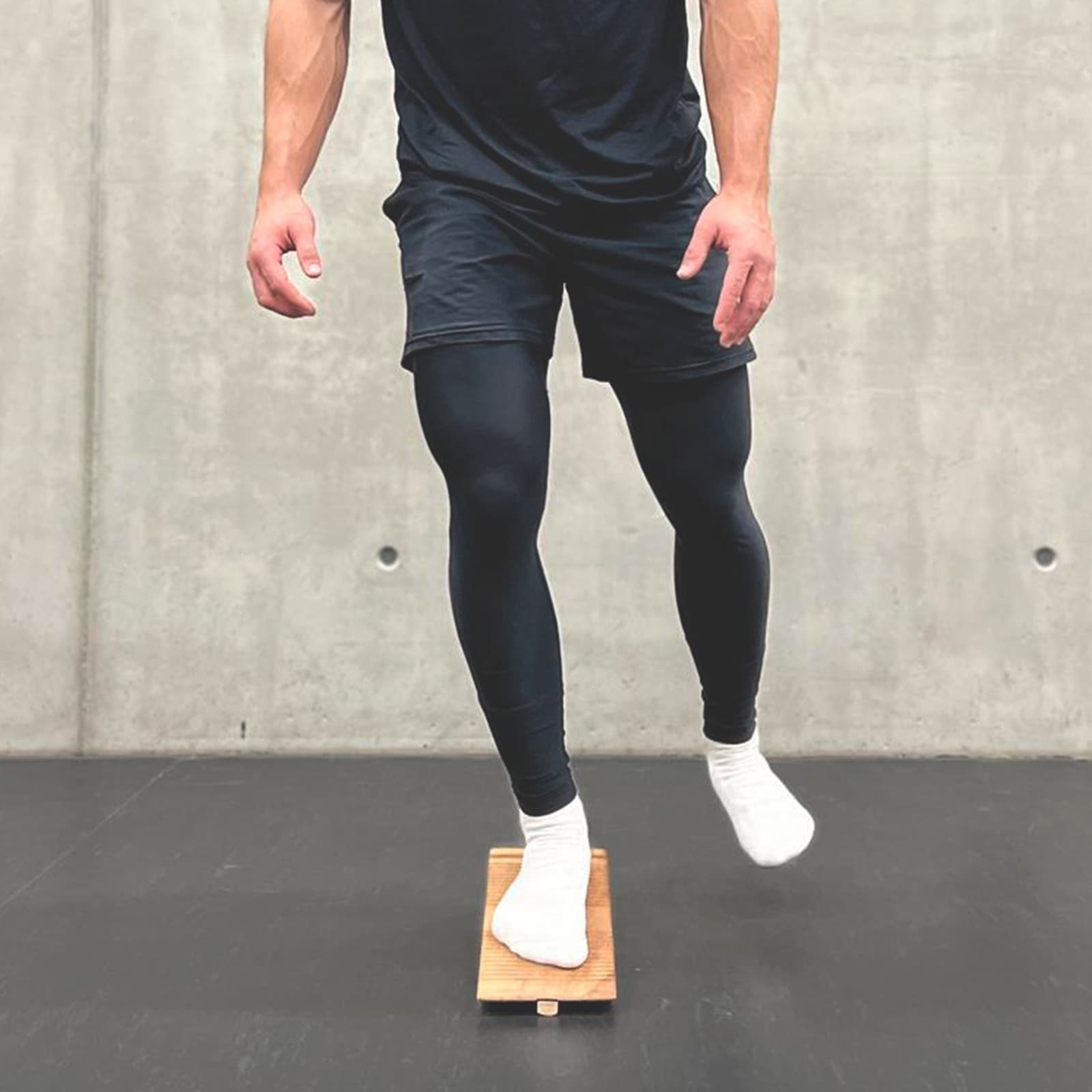 Ein Mann in Sportkleidung trainiert auf Socken mit dem rollholz Balancekeil aus Holz entlang der Längsachse zur Mobilisierung des Fußes vor einer Betonwand.