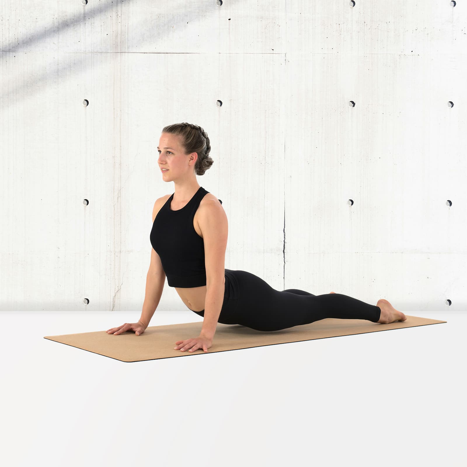 Eine Frau in schwarzer Sportkleidung führt auf der rollholz Korkmatte eine Kobra-Yoga-Übung (Rückbeuge) zur Dehnung des Bauchs und der vorderen Kette aus, vor einer hellen Sichtbetonwand.