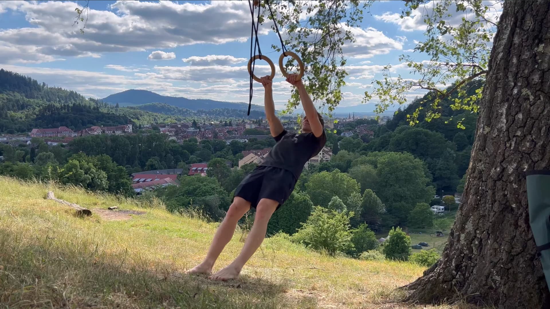 Ein Mann trainiert barfuß auf einer Wiese mit dem rollholz Schlingentrainer aus Holz, der an einem Baum aufgehängt ist, mit Blick auf Freiburg.